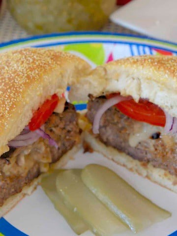 Juicy burgers filled with melted cheese, caramelized onion and bacon, on a bun on a summery plate with dill pickle slices.